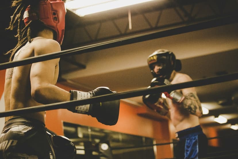 Two boxers wearing headgear and gloves spar inside a ring, one with dreadlocks and the other with arm tattoos.