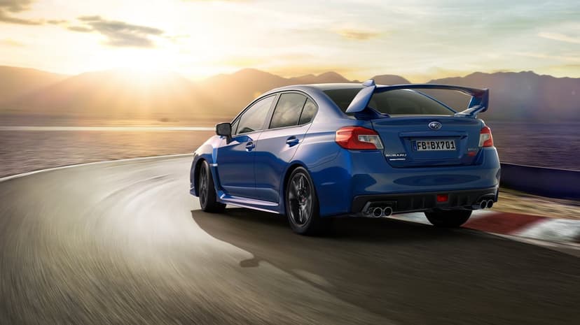 Blue Subaru WRX STI sports car driving on a scenic coastal road with mountains during a golden sunset.