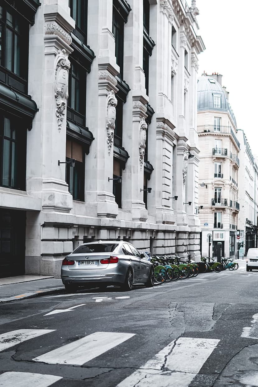 Silver BMW sedan parked beside green rental bikes against ornate Parisian architecture with classical columns.