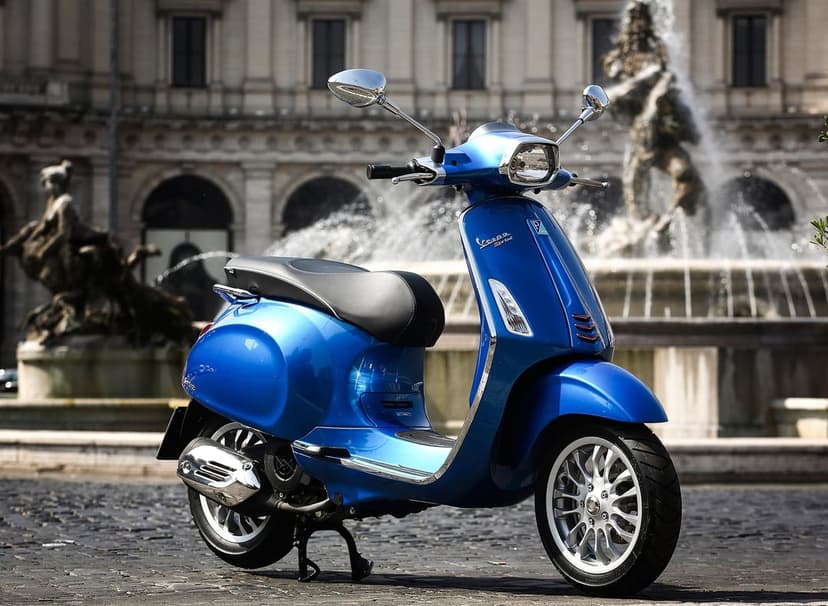 Blue Vespa Sprint scooter on cobblestones with the Fountain of the Naiads and classical Roman buildings blurred behind.