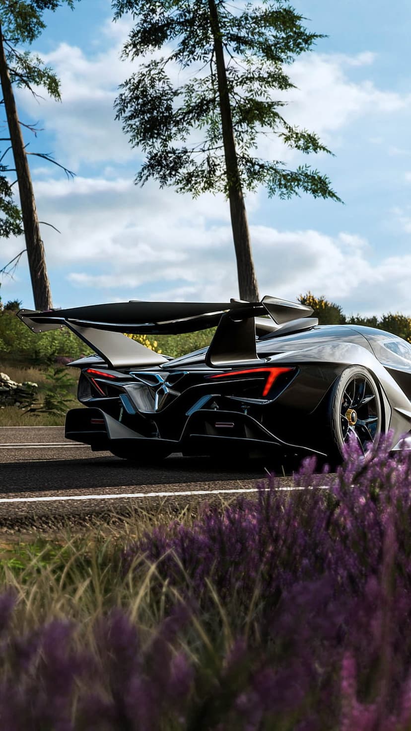 Black sports car with a large rear wing drives past purple wildflowers on a winding road through a tall forest.