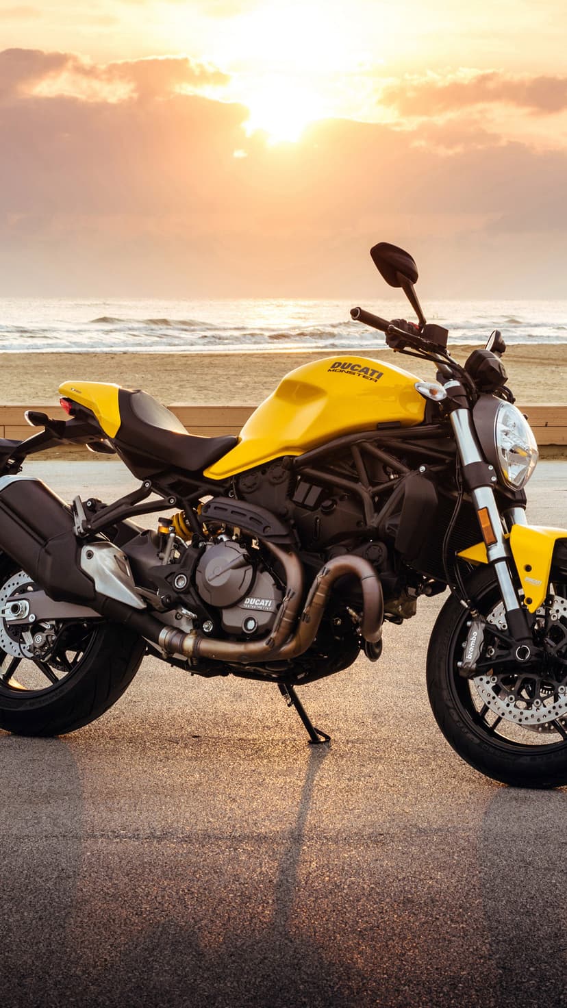 Yellow Ducati Monster motorcycle parked on wet asphalt during a golden sunset with cloudy ocean views.