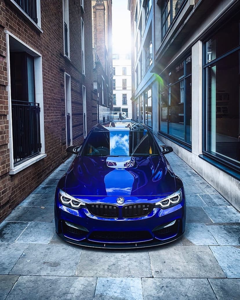 Blue BMW M4 sports car parked in a sunlit London alleyway between historic brick walls and modern glass facades.