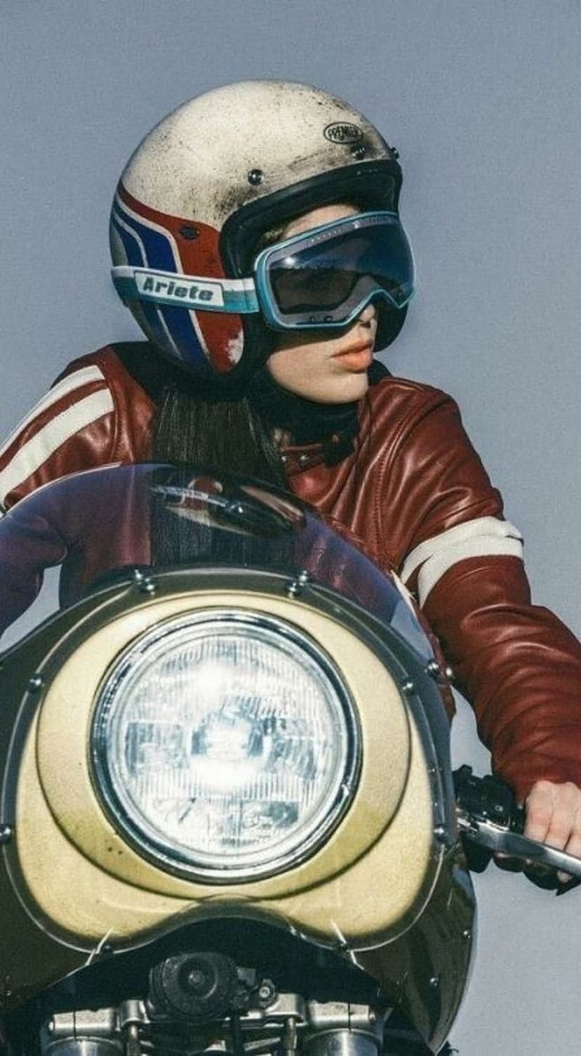 Woman in a red leather jacket and striped helmet sits on a vintage cafe racer motorcycle with goggles.