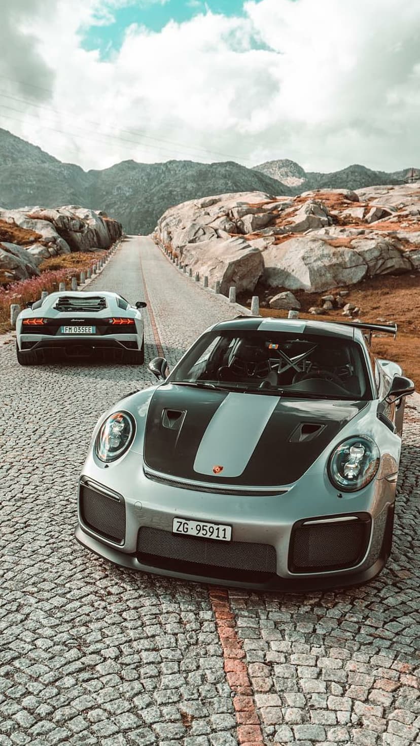 Silver Porsche GT2 RS and white Lamborghini Aventador parked on a cobblestone mountain road under cloudy skies.