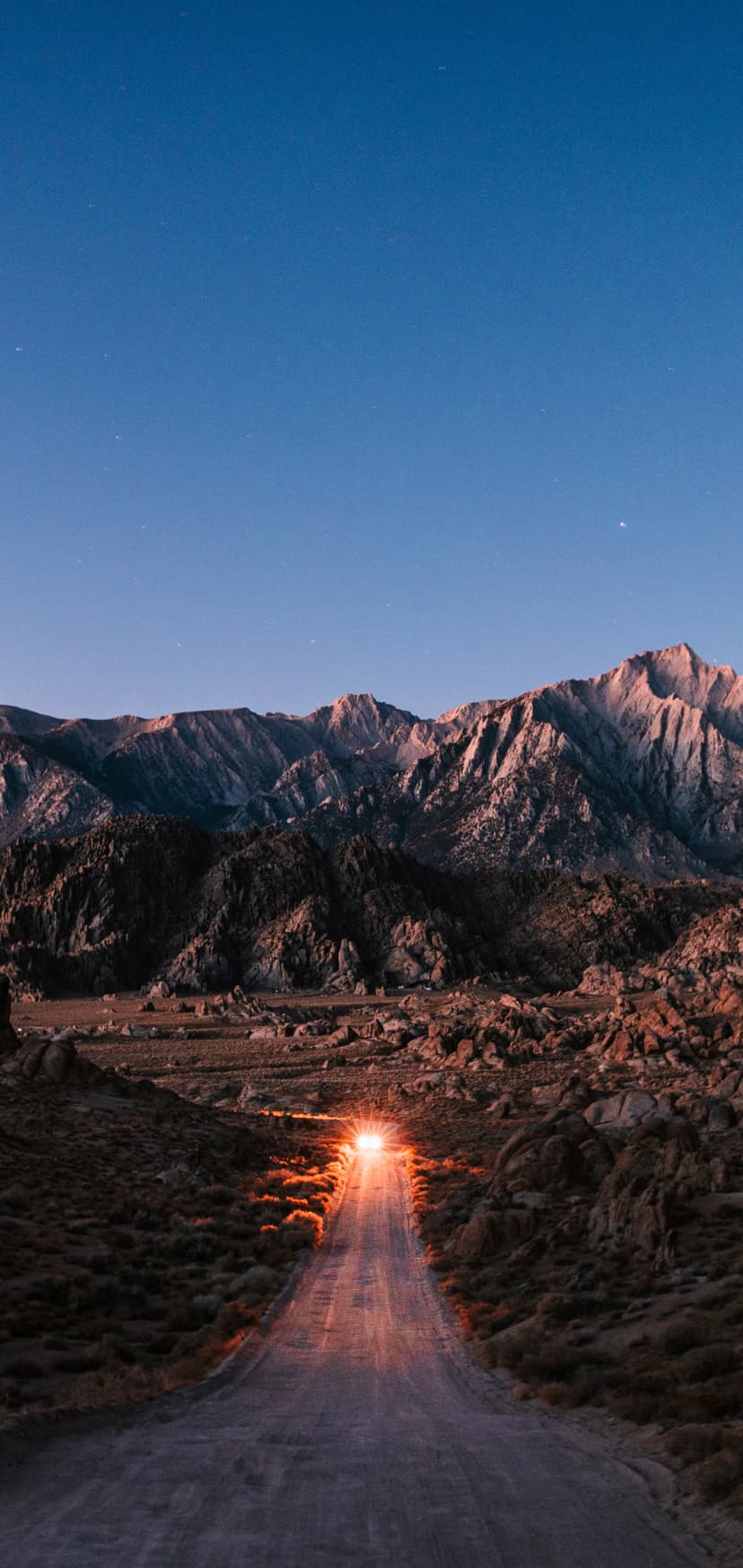 Dirt road glowing with orange light leads toward distant mountains under a starry twilight sky in the desert.