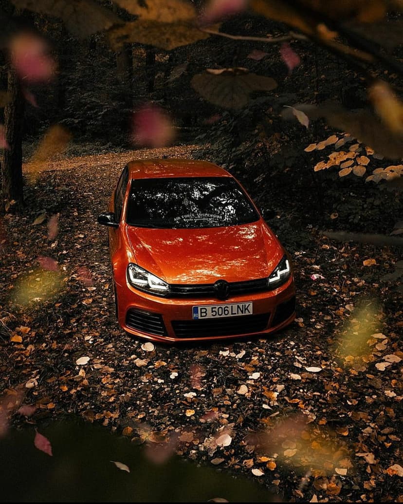 Orange Volkswagen Golf MK6 with glowing headlights parked on a leaf-strewn forest path during a misty autumn day.