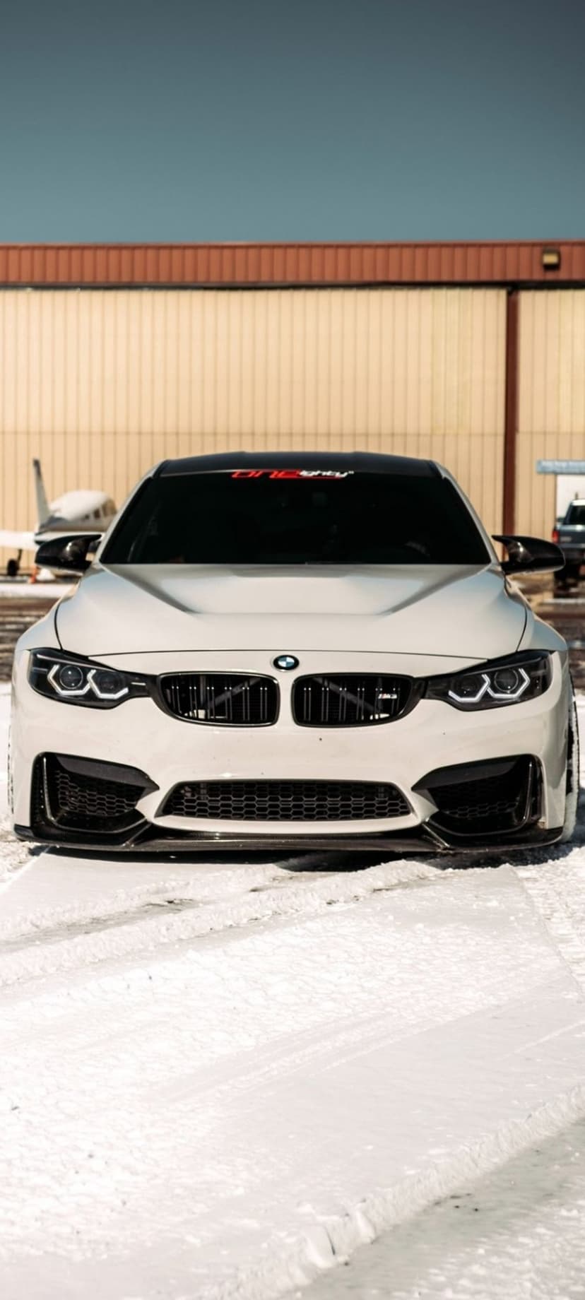 White BMW M4 coupe parked on snowy ground inside an airport hangar with a sleek private jet in the background.