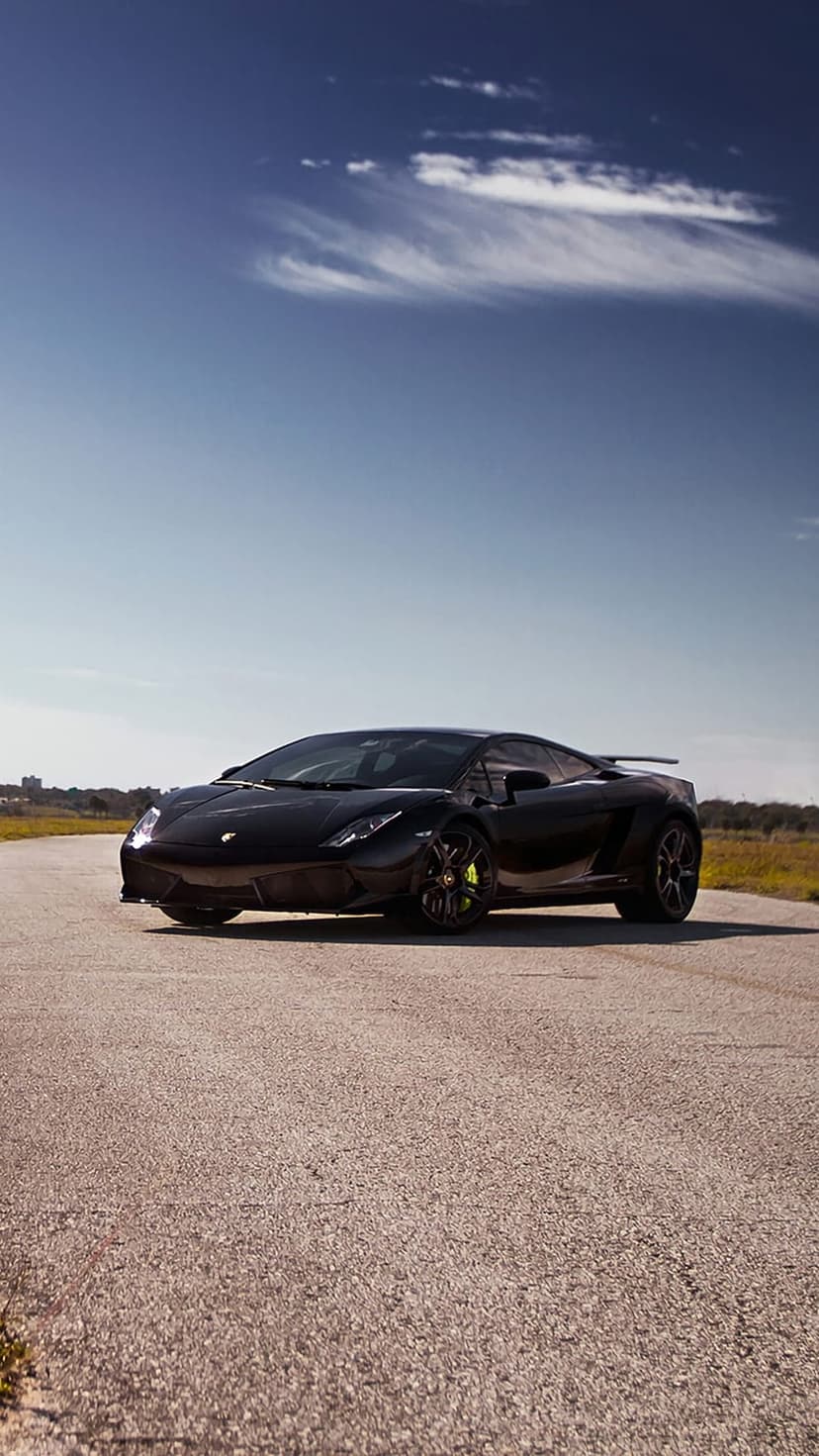 Black Lamborghini Gallardo with yellow brake calipers on a grey asphalt road under a bright blue sky.