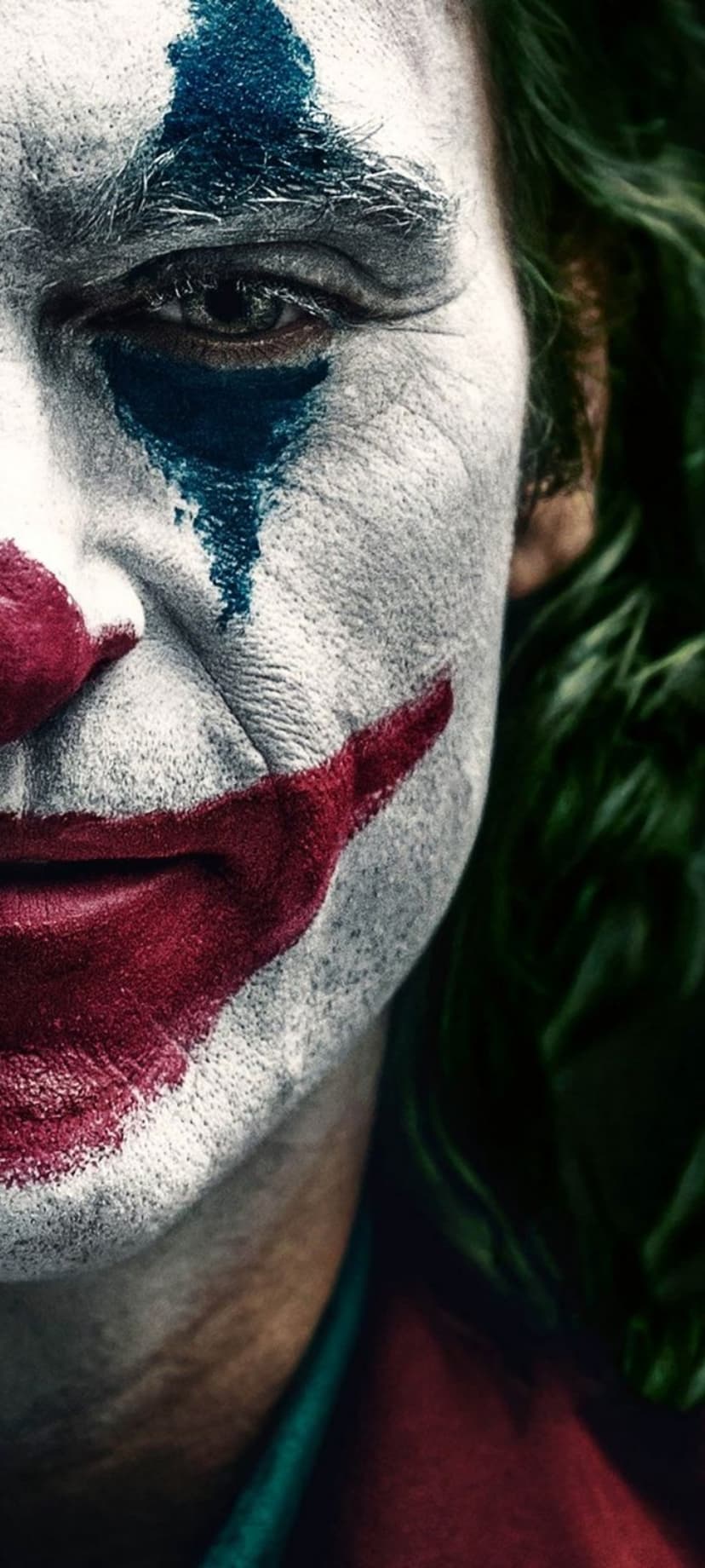 Joaquin Phoenix as Joker shows smeared blue eye paint, white face makeup, and a red smile in a tight close-up.