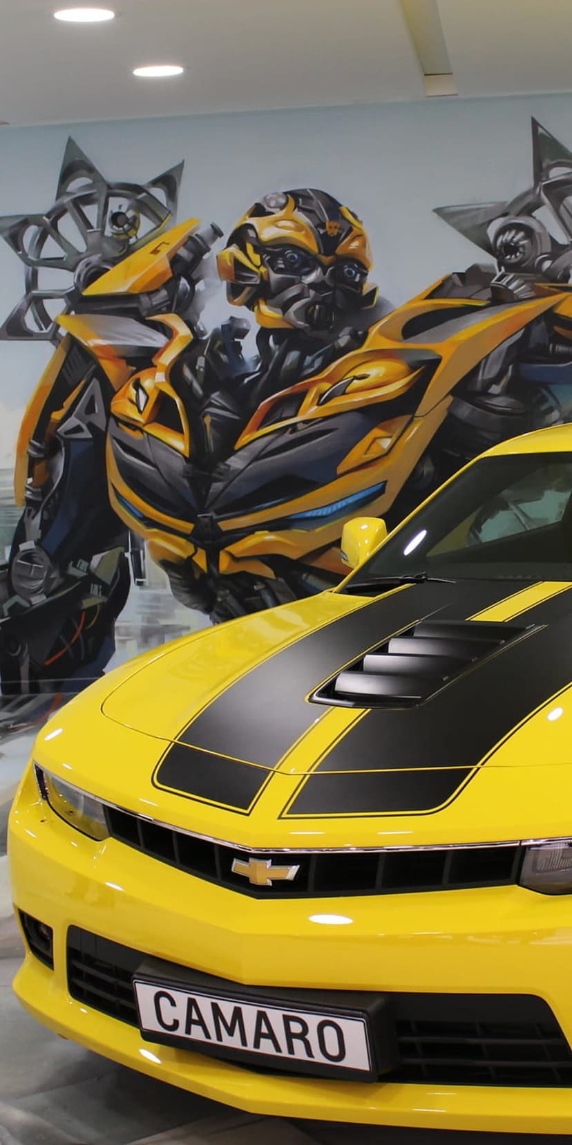 Yellow Chevrolet Camaro with black stripes parked in front of a giant Bumblebee Autobot mural on a brick wall.