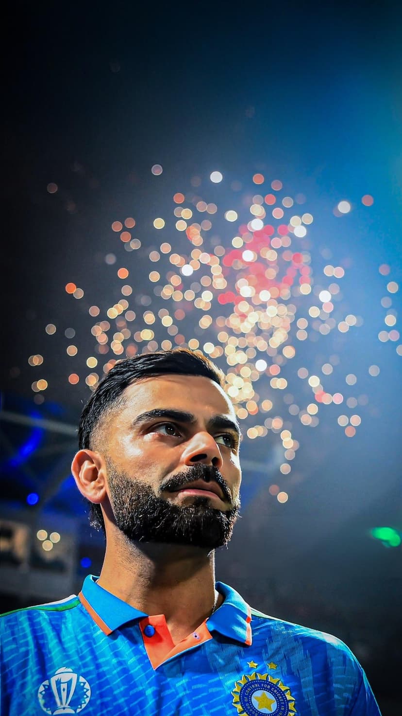 Virat Kohli in a blue jersey gazes upward against a background of red and white bokeh celebratory lights.