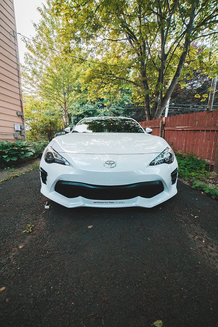 White Toyota 86 sports car parked on a dark driveway with green trees and a wooden fence in the background.