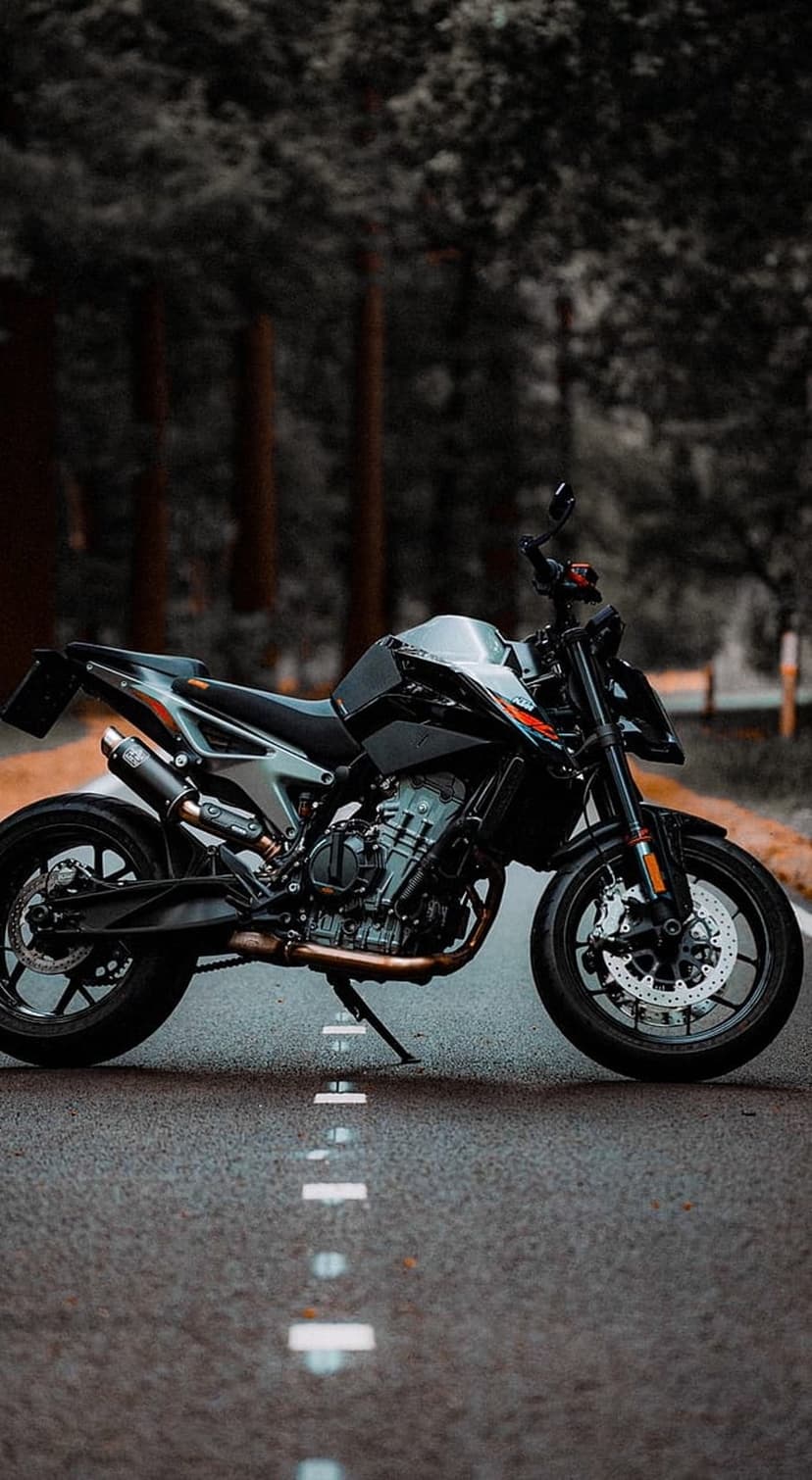 KTM motorcycle parked on an asphalt forest road with tall trees and dramatic low-light metallic details.