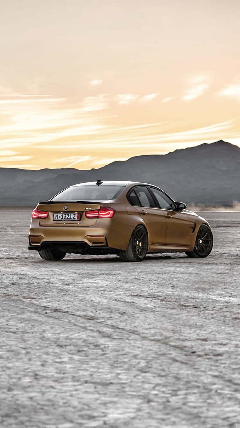 Golden BMW M3 sedan drifting through desert sand dunes during sunset with a mountain range in the background.