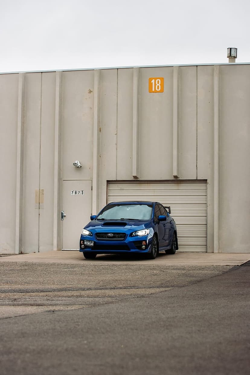 Blue Subaru WRX with illuminated headlights parked on asphalt in front of a beige industrial garage building.