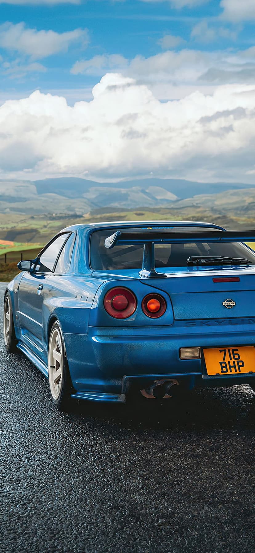 Blue Nissan Skyline R34 GTR with a large rear wing parked on a wet road against green hills and a cloudy sky.