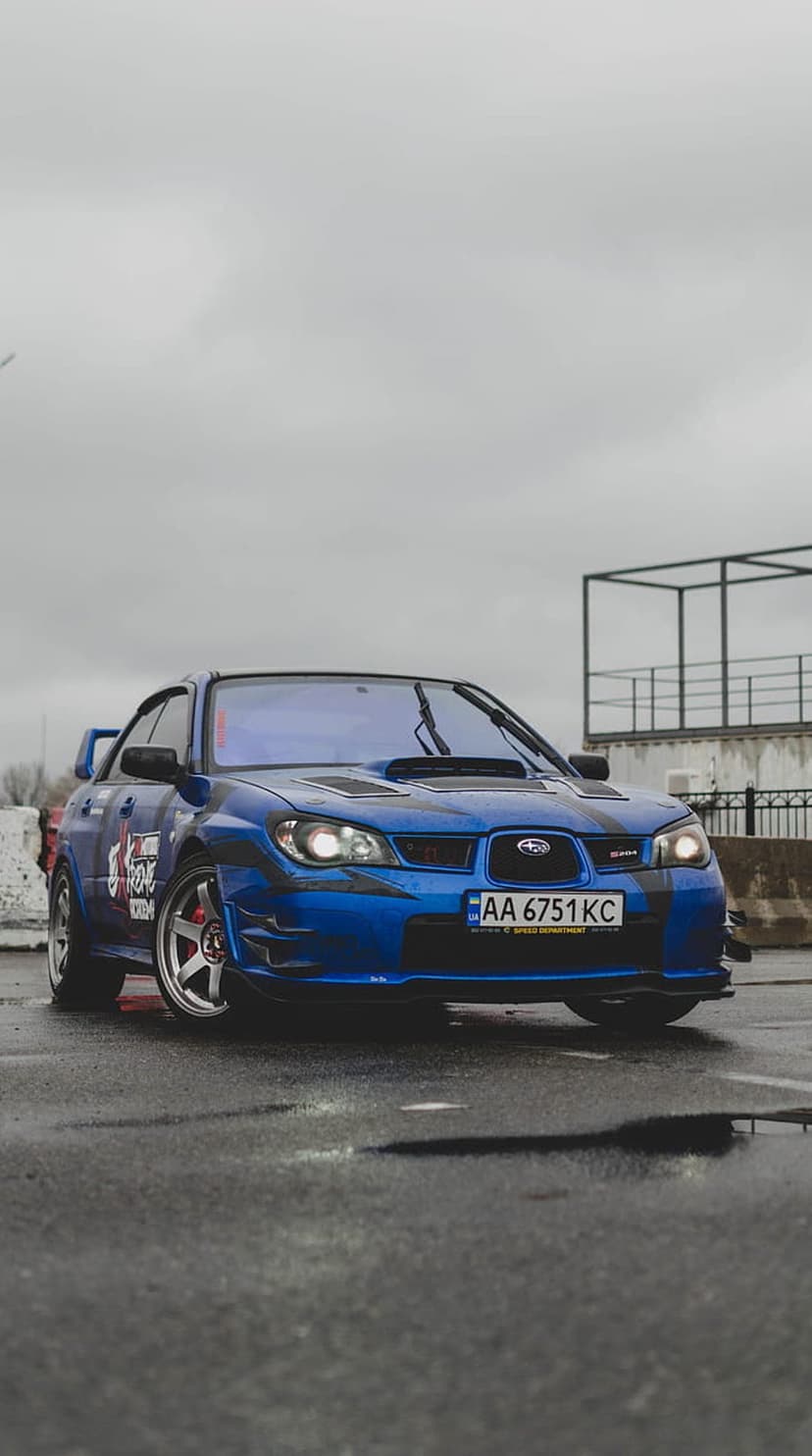 Blue Subaru Impreza WRX STI with racing decals on wet urban asphalt under a moody and overcast city sky.