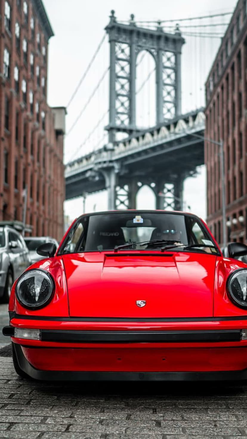 Red Porsche 911 classic car parked on cobblestone street with Brooklyn Bridge and brick buildings behind it.