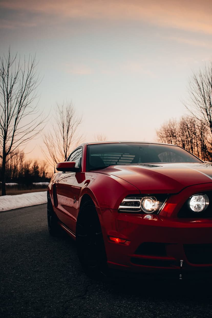 Red Ford Mustang with glowing headlights parked on a dark road beside snow under a pink and orange sunset sky.