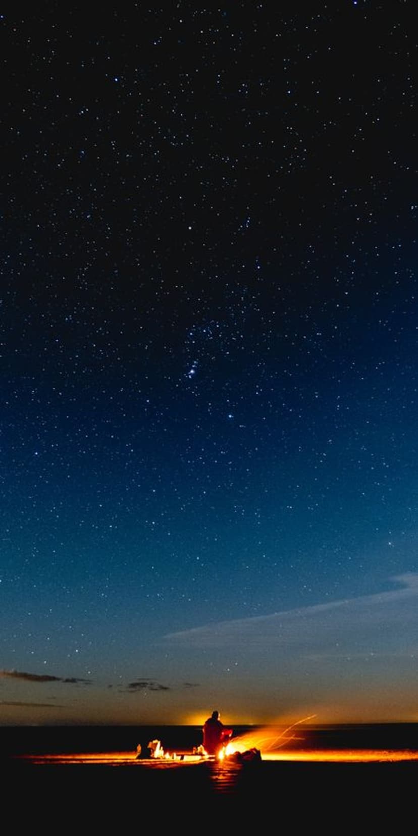 Person sitting by a campfire on a riverbank under a vast starry sky with a purple and black celestial gradient.