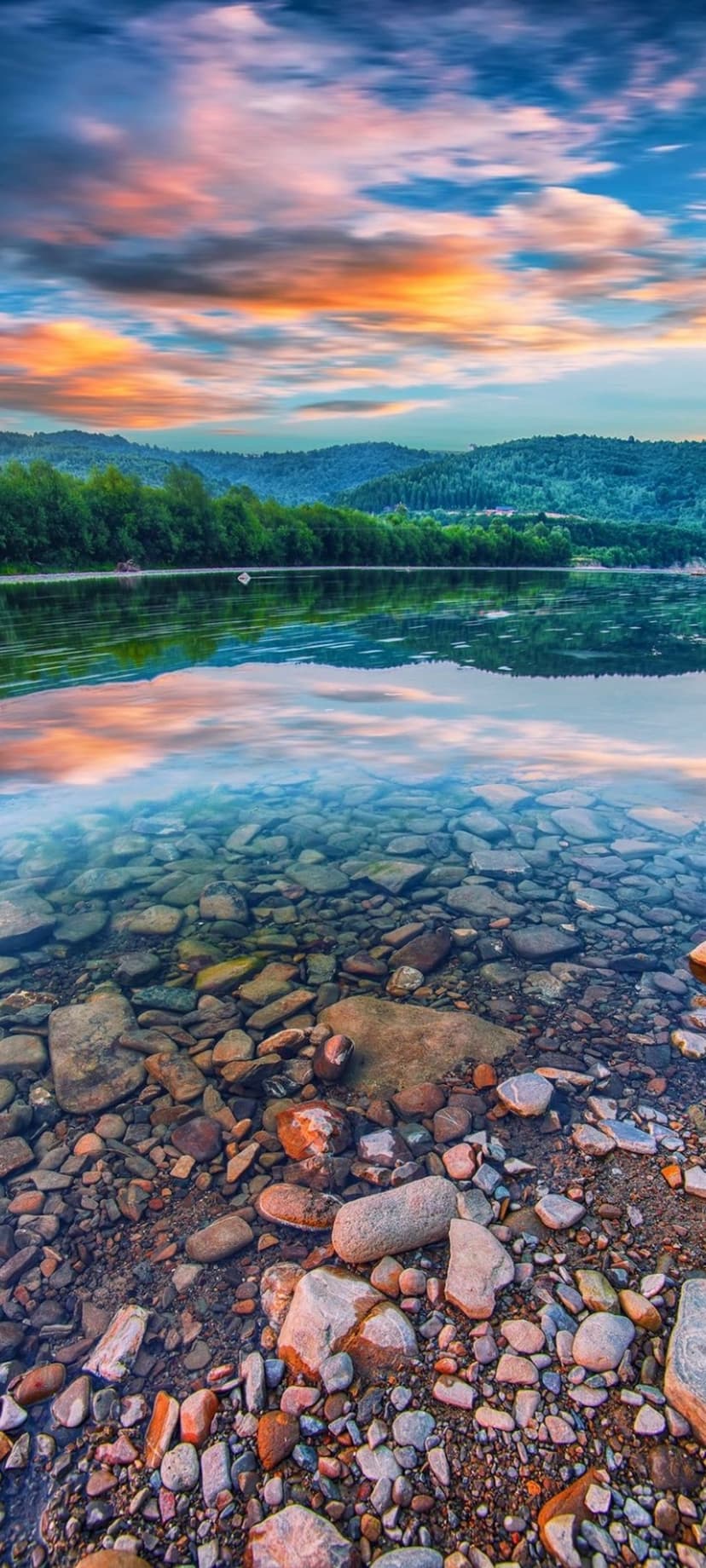 Rocky lake shore with clear water reflecting a pink and orange sunset sky behind silhouetted forest hills.