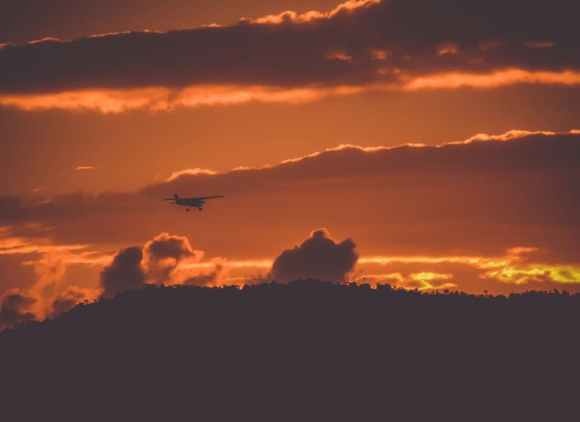 Small airplane silhouetted against a radiant orange sunset sky with dark rolling hills in the background.