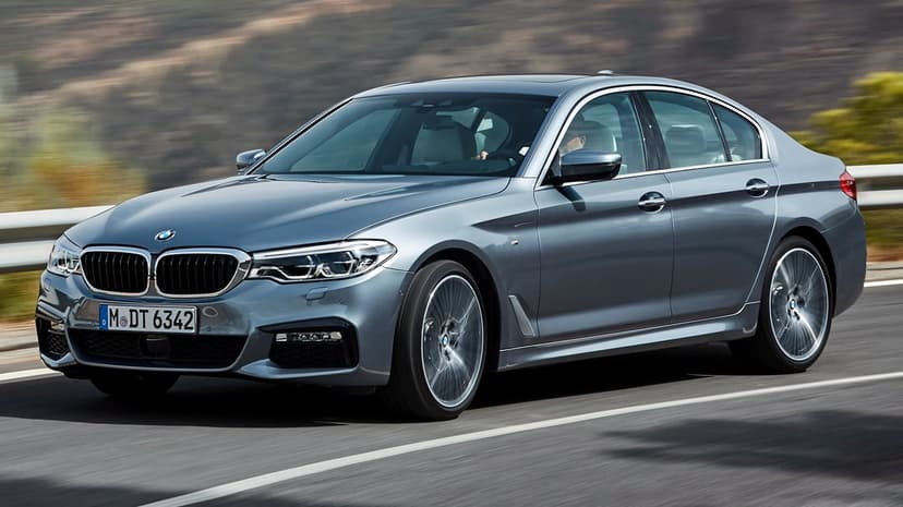 Grey BMW 5 Series M Sport sedan driving on a highway with motion-blurred wheels and background landscape.