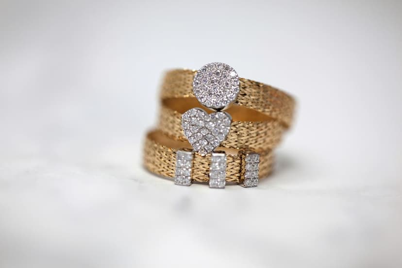 Diamond heart and circle rings on a woven gold mesh band sparkle under soft lighting with intricate jewelry detail.