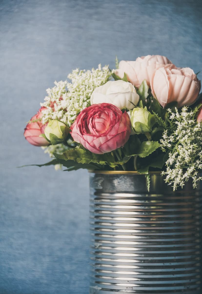 Pink and white ranunculus flowers in a corrugated tin can vase against a textured light blue background.