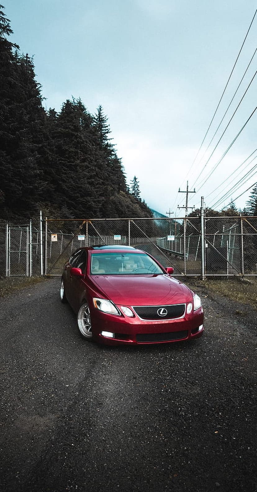 Red Lexus GS luxury sedan parked on a gravel road before a gated pine forest under a moody overcast sky.
