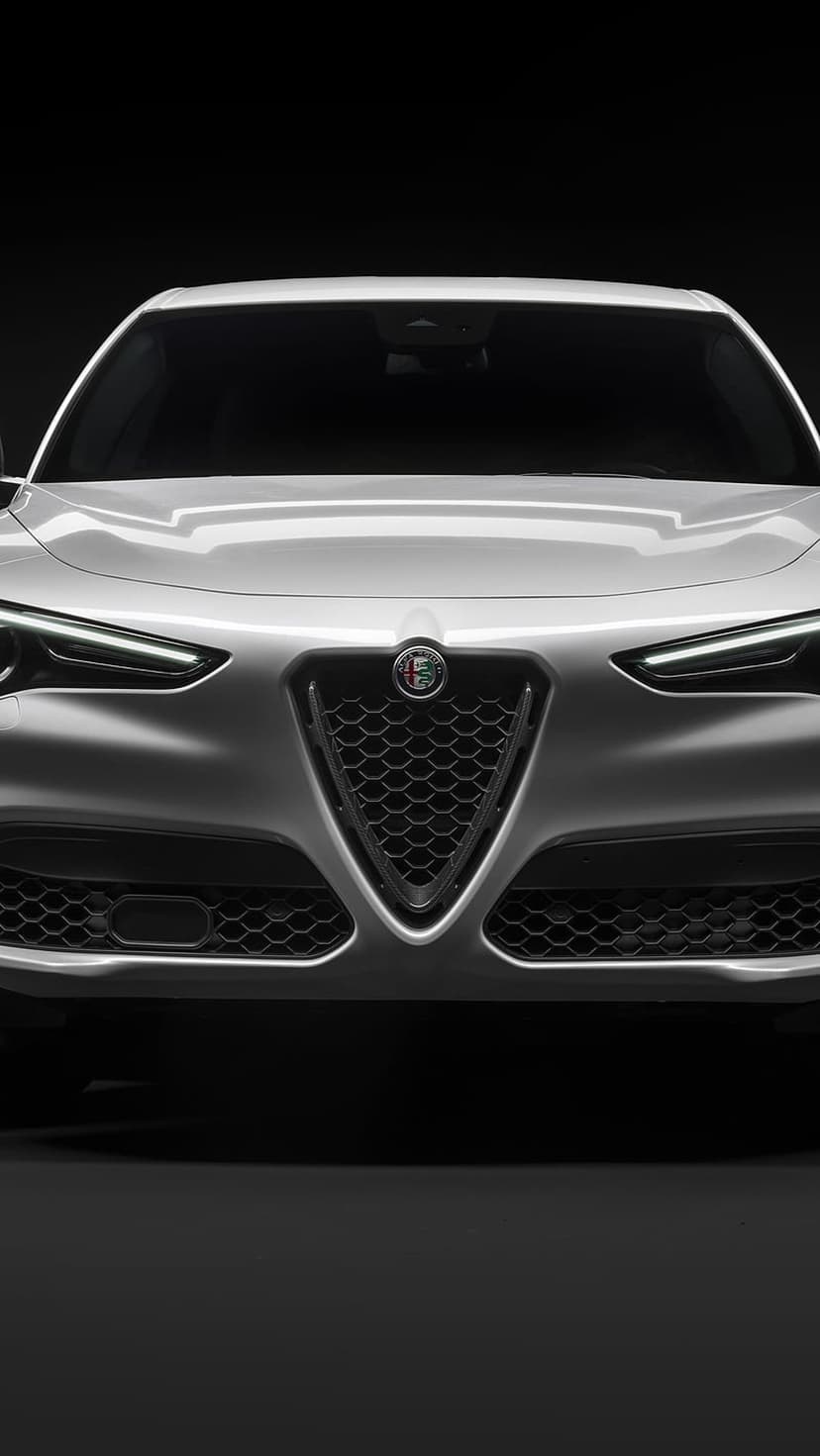 White Alfa Romeo Stelvio SUV front view with illuminated LED headlights and shield grille on a black background.