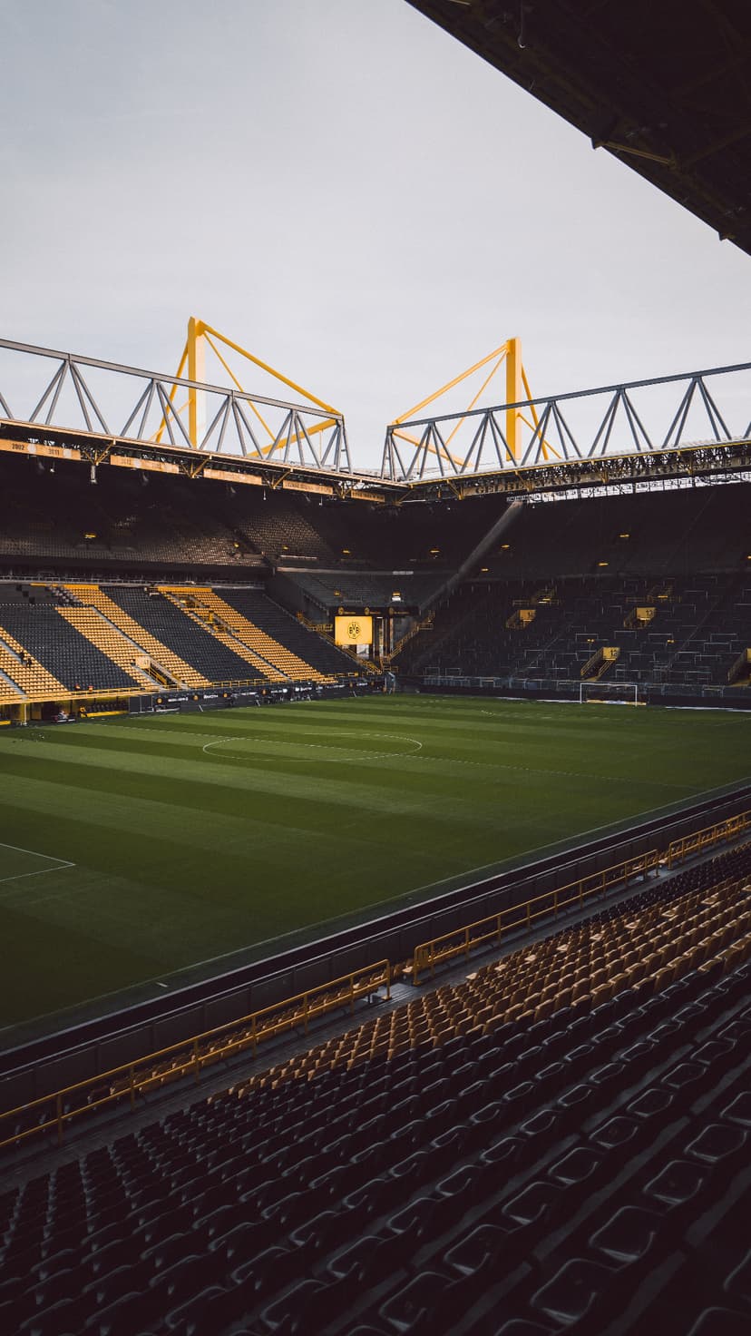 Yellow and black stadium seats overlook a striped green football pitch under industrial yellow roof structures.
