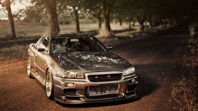 Silver Nissan Skyline GT-R R34 with ARC intercooler on a tree-lined dirt path during warm golden hour light.