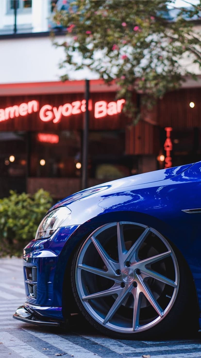 Blue sports car with silver rims parked on cobblestones next to a red neon Ramen Gyoza Bar sign at night.