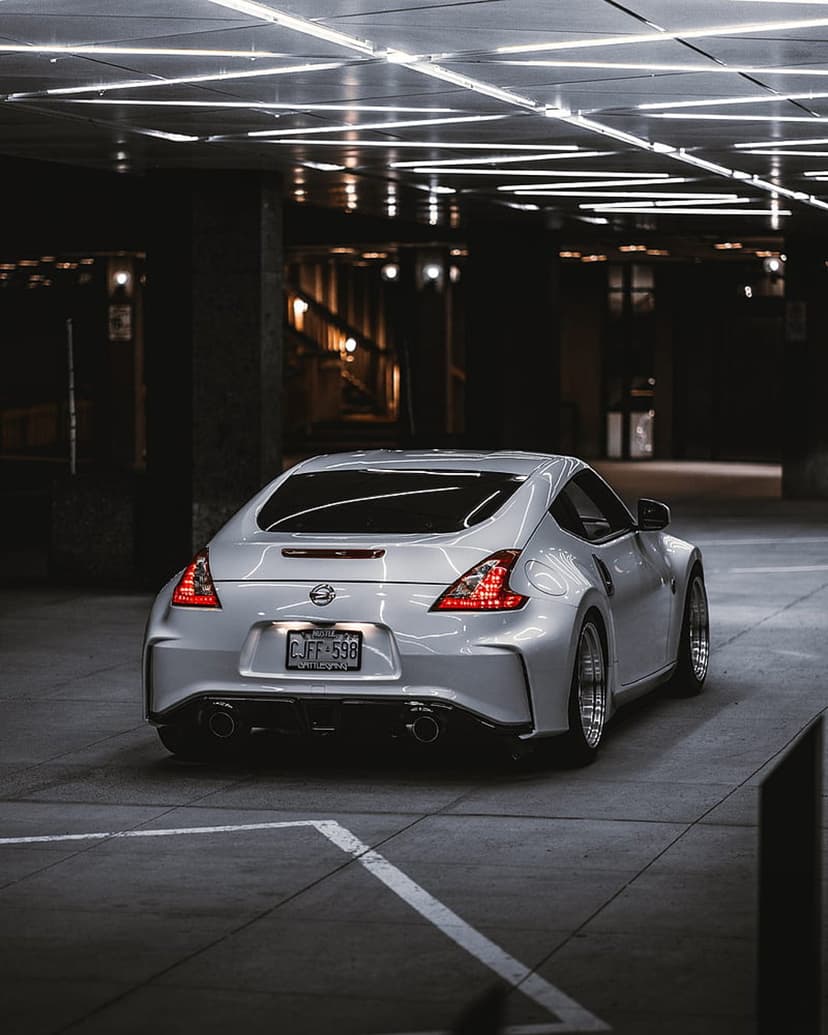 Nissan 370Z rear view with lit red taillights parked under linear ceiling lamps in a dark concrete parking garage.