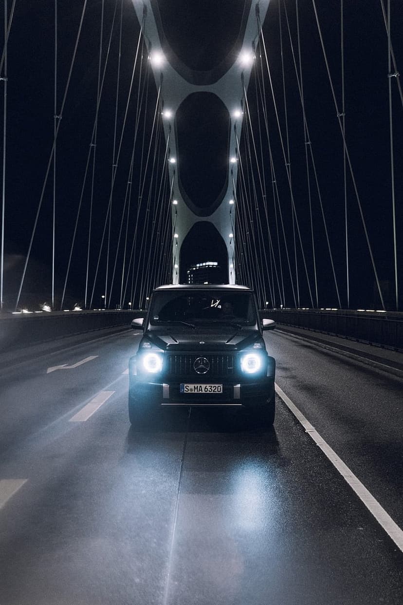 Mercedes-AMG G 63 SUV driving on a wet bridge at night under illuminated cables with bright headlights glowing.
