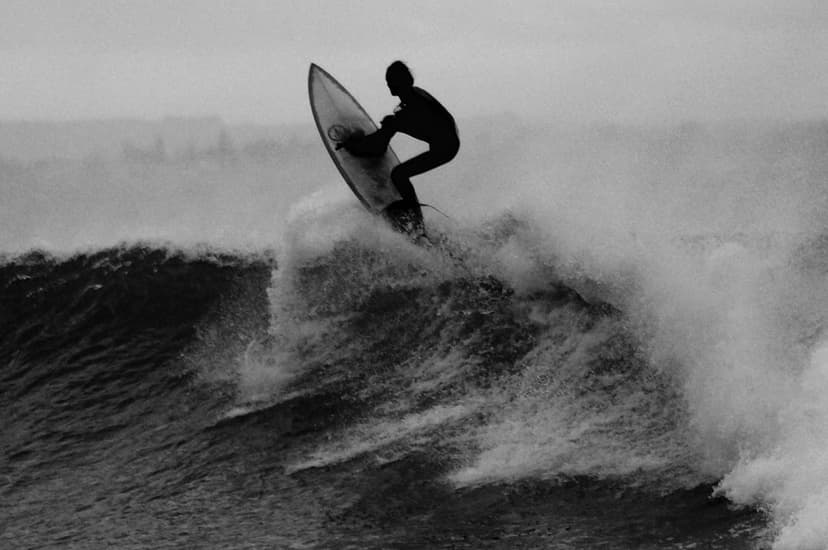 Surfer silhouetted against a cresting ocean wave with dramatic white sea foam and a hazy gray horizon line.
