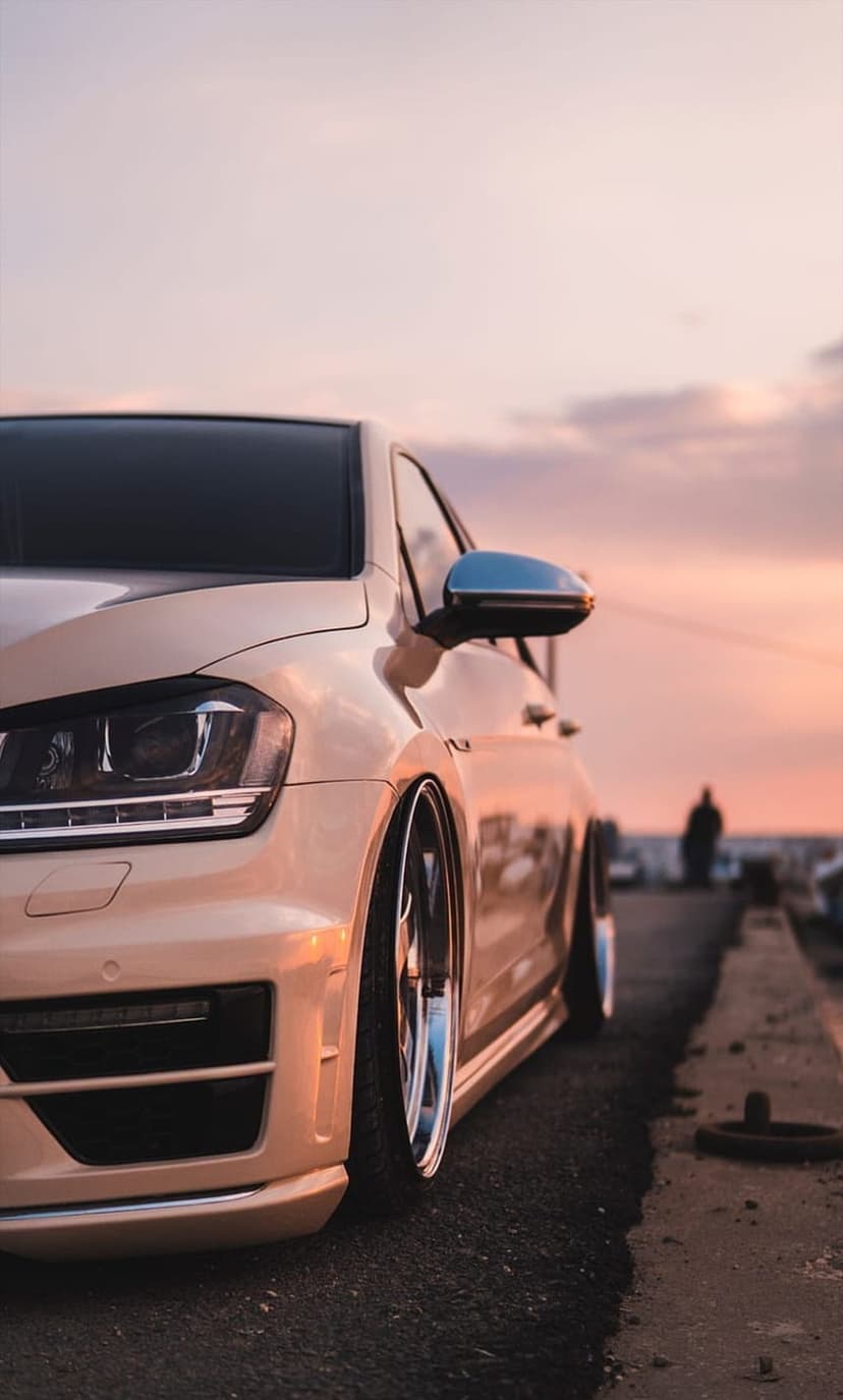 Beige Volkswagen Golf R with chrome wheels parked on a coastal road during a golden sunset by the beach.