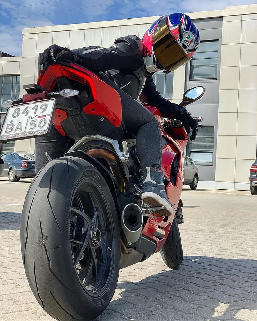 Rider in red and black gear sits on a red Ducati motorcycle with a gold visor against a modern building.