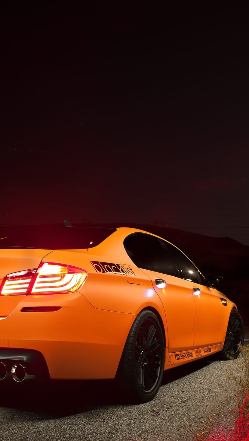 BMW M5 sedan with matte orange finish and black rims parked on a dark street with glowing red taillights.