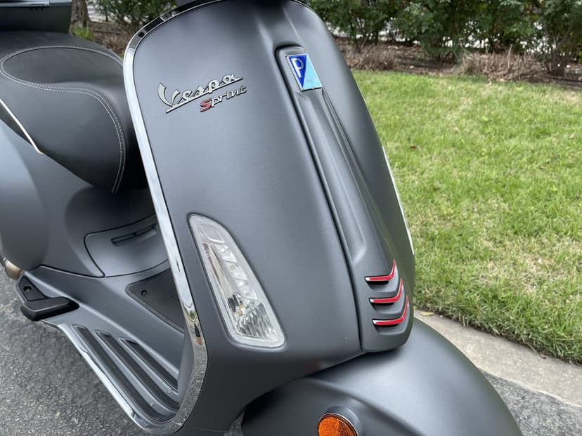 Matte grey Vespa Sprint scooter front shield with red accents and chrome logo on a sunny day.