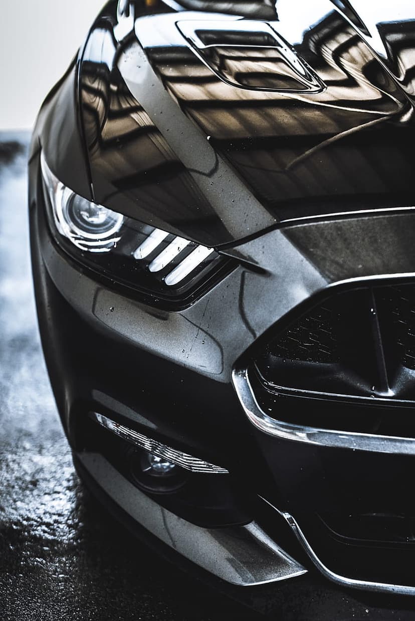 Black Ford Mustang LED headlight and chrome grille with water droplets and architecture reflections.