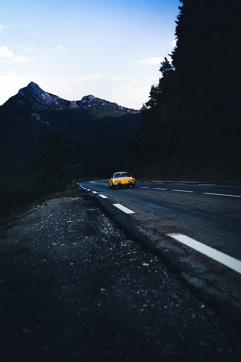 Yellow sports car driving on a winding asphalt road through a dark pine forest with a jagged mountain peak at dusk.