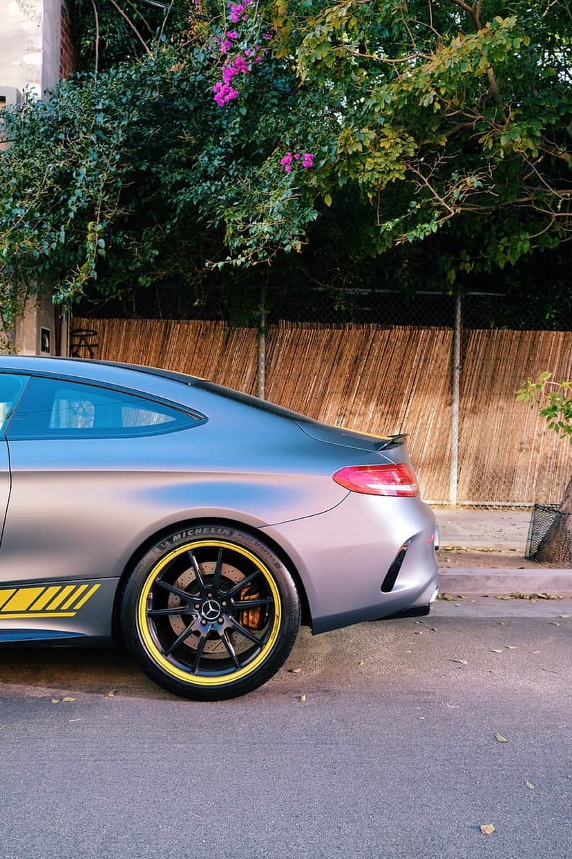 Matte grey Mercedes coupe with yellow racing stripes on the side skirt and wheels parked next to a bamboo fence.