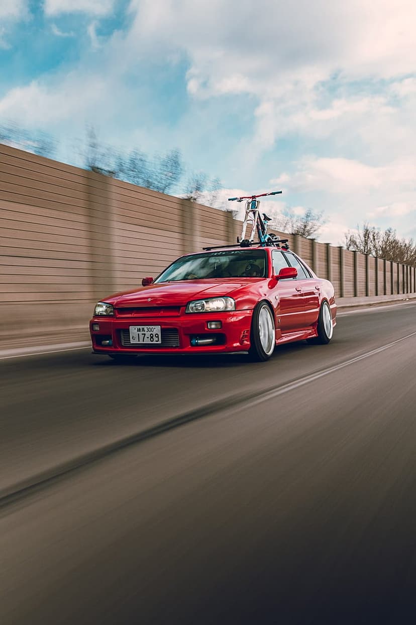 Red Nissan Skyline R34 sedan with a bicycle on its roof rack driving past a blurred highway sound barrier.