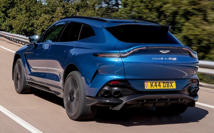 Blue Aston Martin DBX707 SUV driving on a sunlit road with green trees and motion blur in the background.