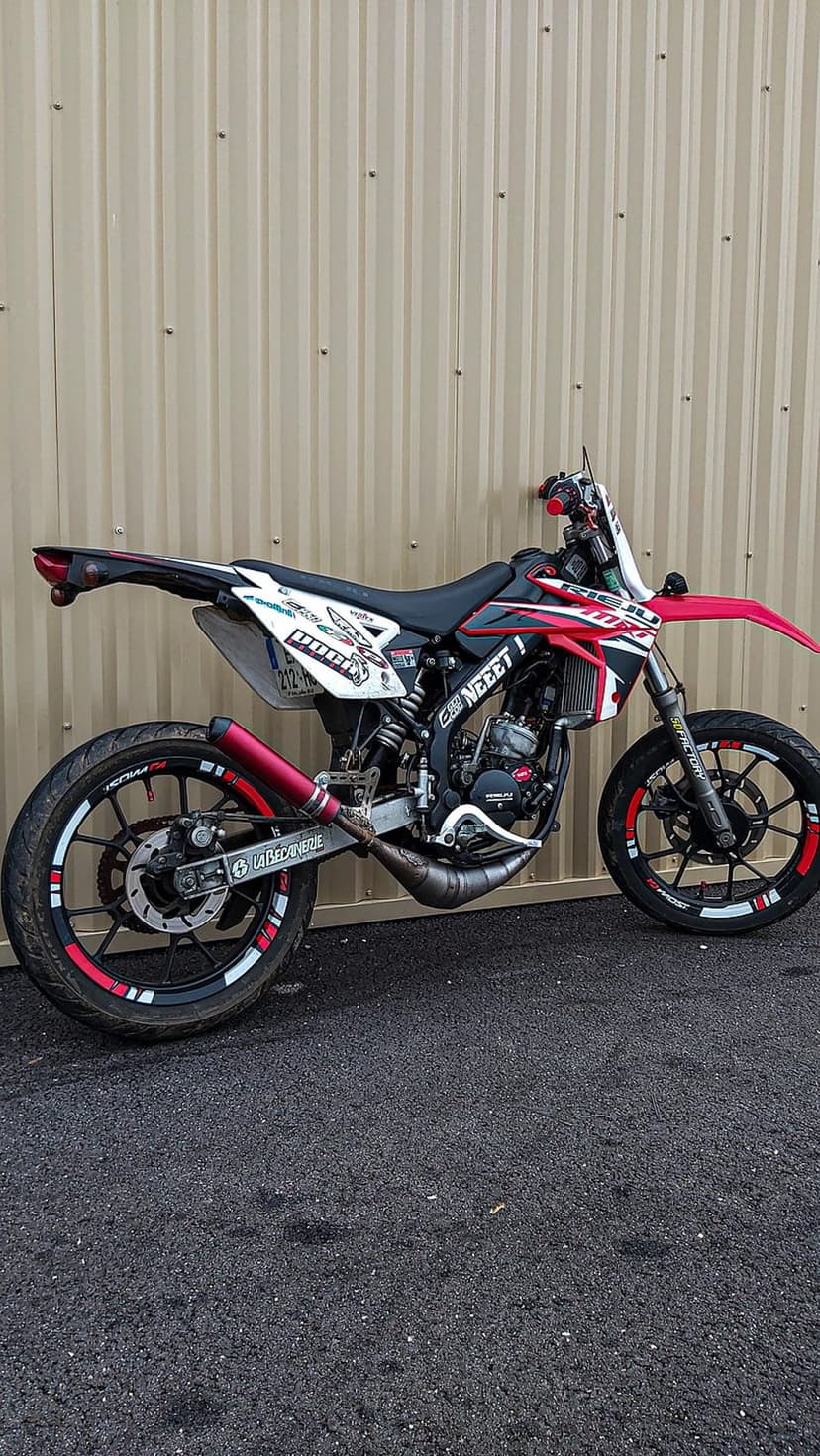 Red and white supermoto motorcycle with custom decals and red rims parked against a corrugated beige metal wall.