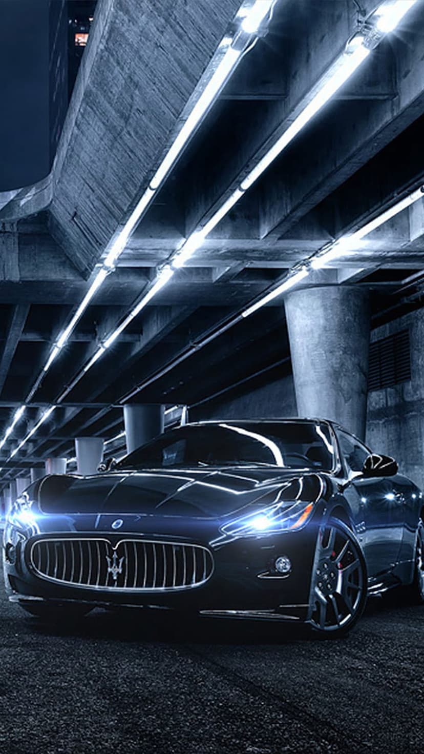 Maserati GranTurismo sports car with glowing blue headlights parked in a dark, brutalist concrete underpass.