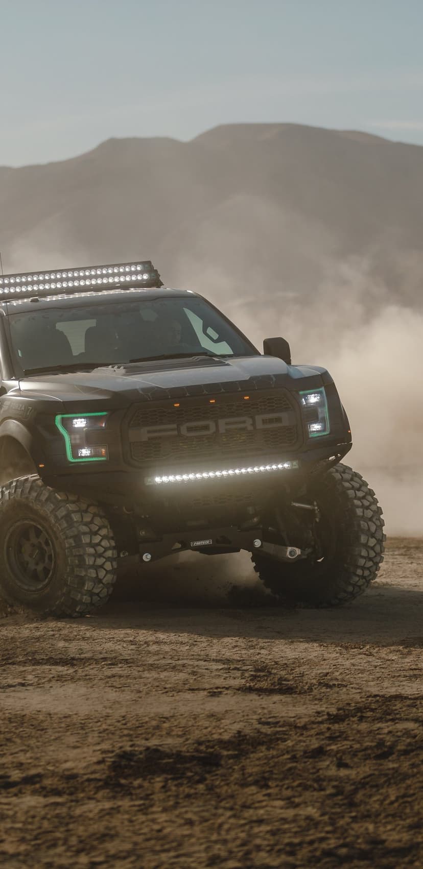 Black Ford Raptor off-road truck with glowing LED bars driving through heavy desert dust and sand clouds.